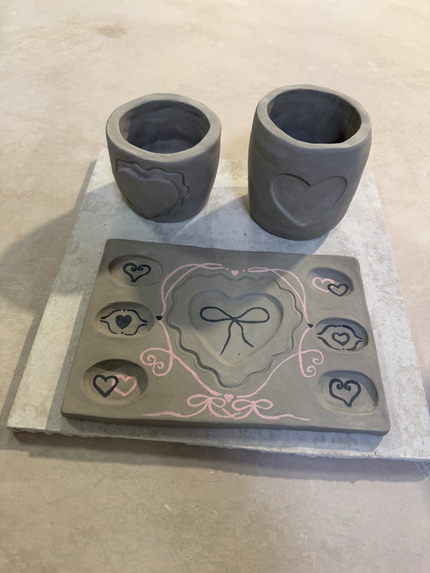 Unfired gray clay cups and a heart palette tray painted with hearts and pink ribbons