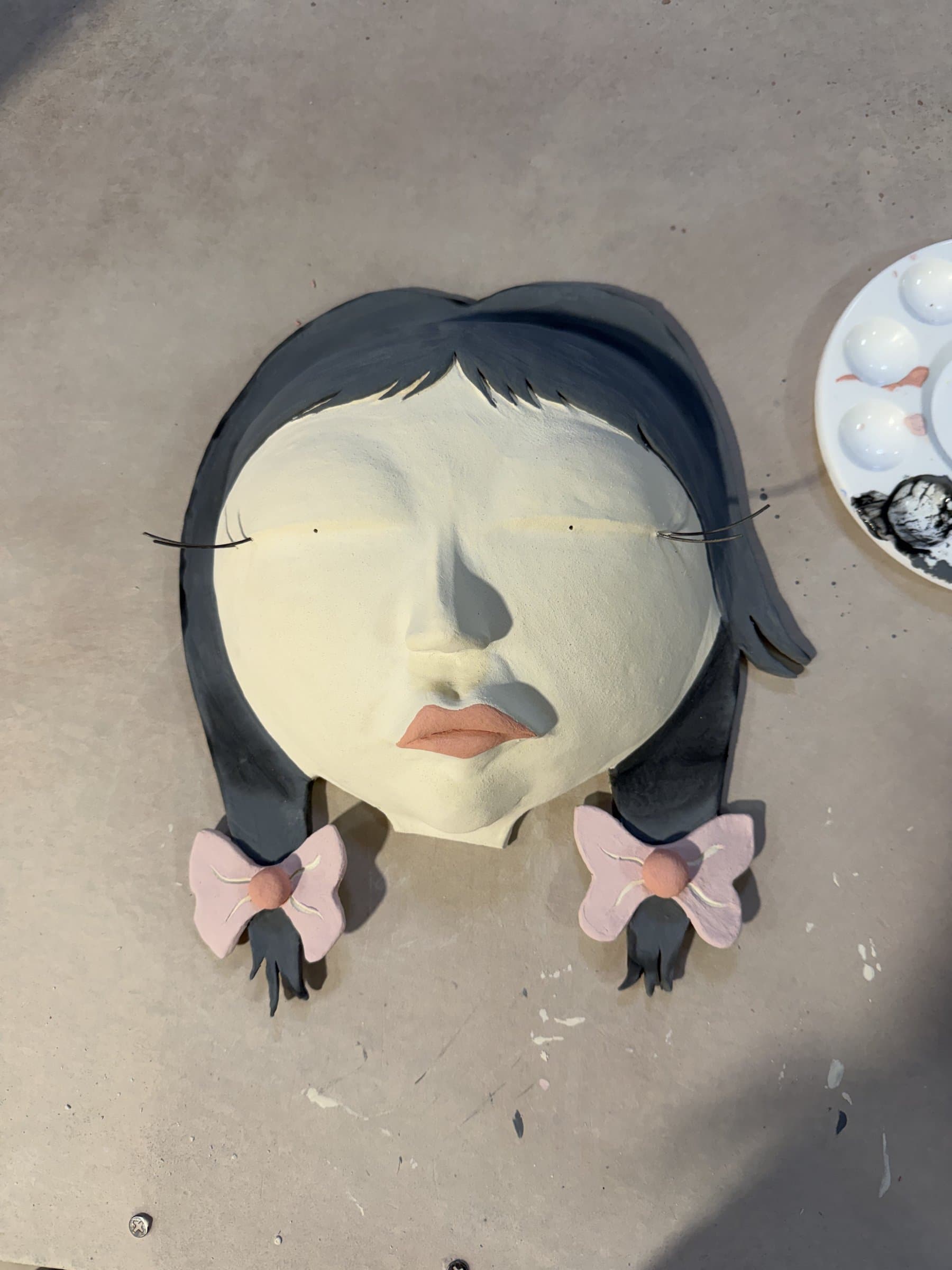 The bisque-fired face painted in soft matte colors with pink bows, beside a paint palette