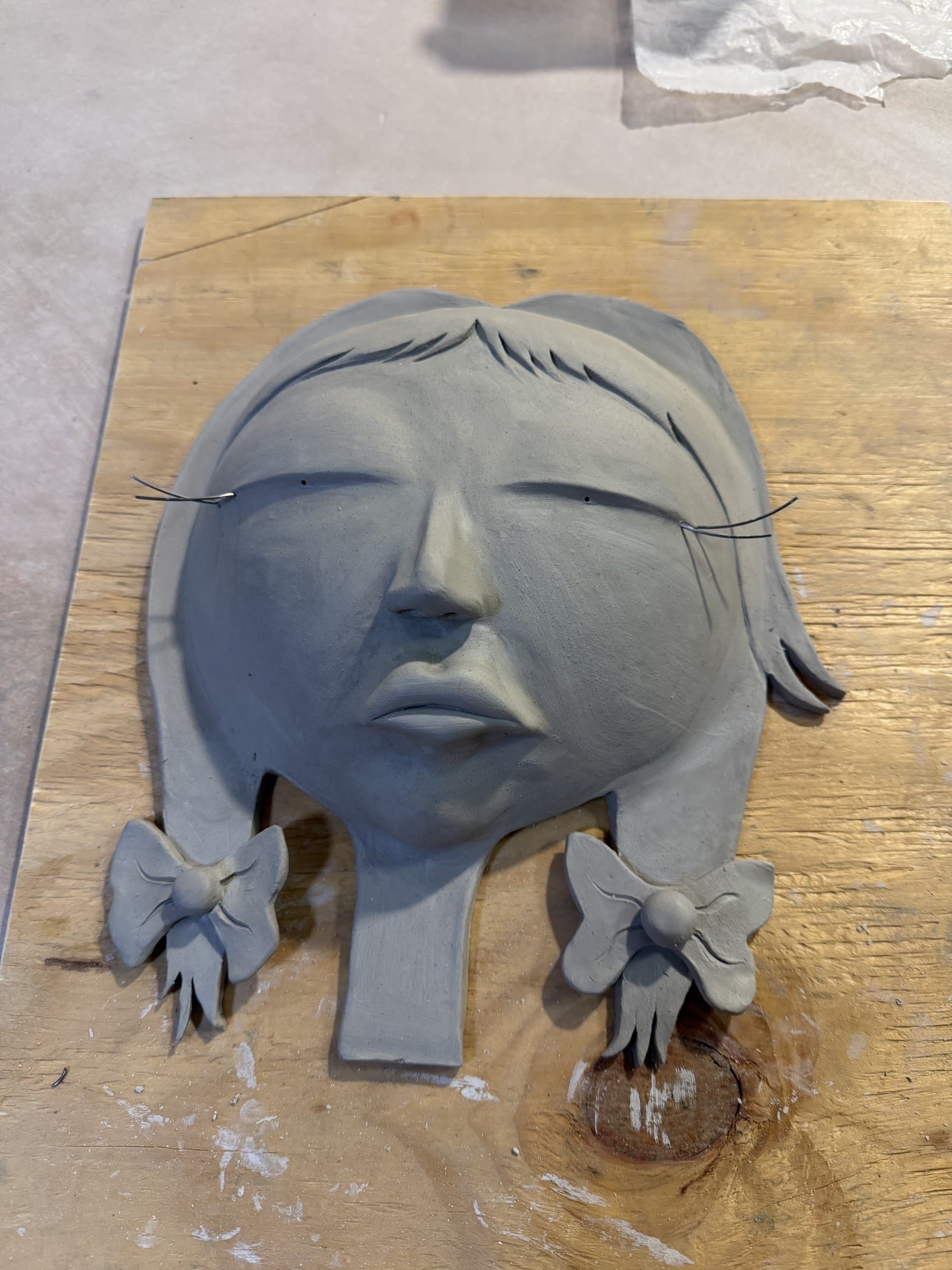 A raw gray clay face sculpture with braids, bows, and wire eyelashes resting on a wood board