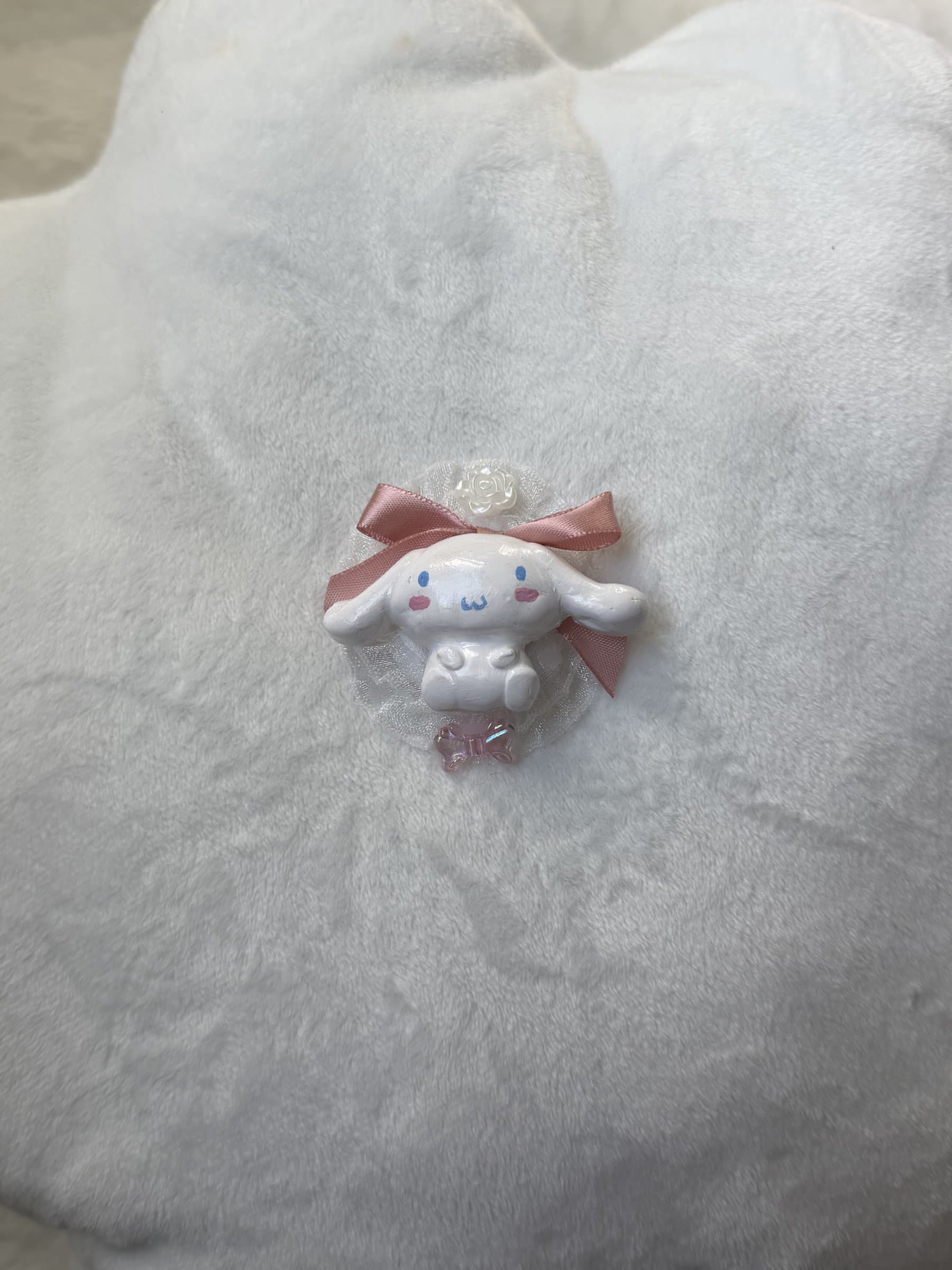 Clay Cinnamoroll in a white lace rosette with a dusty pink satin bow, pearl rose, and pink bow bead, on white plush
