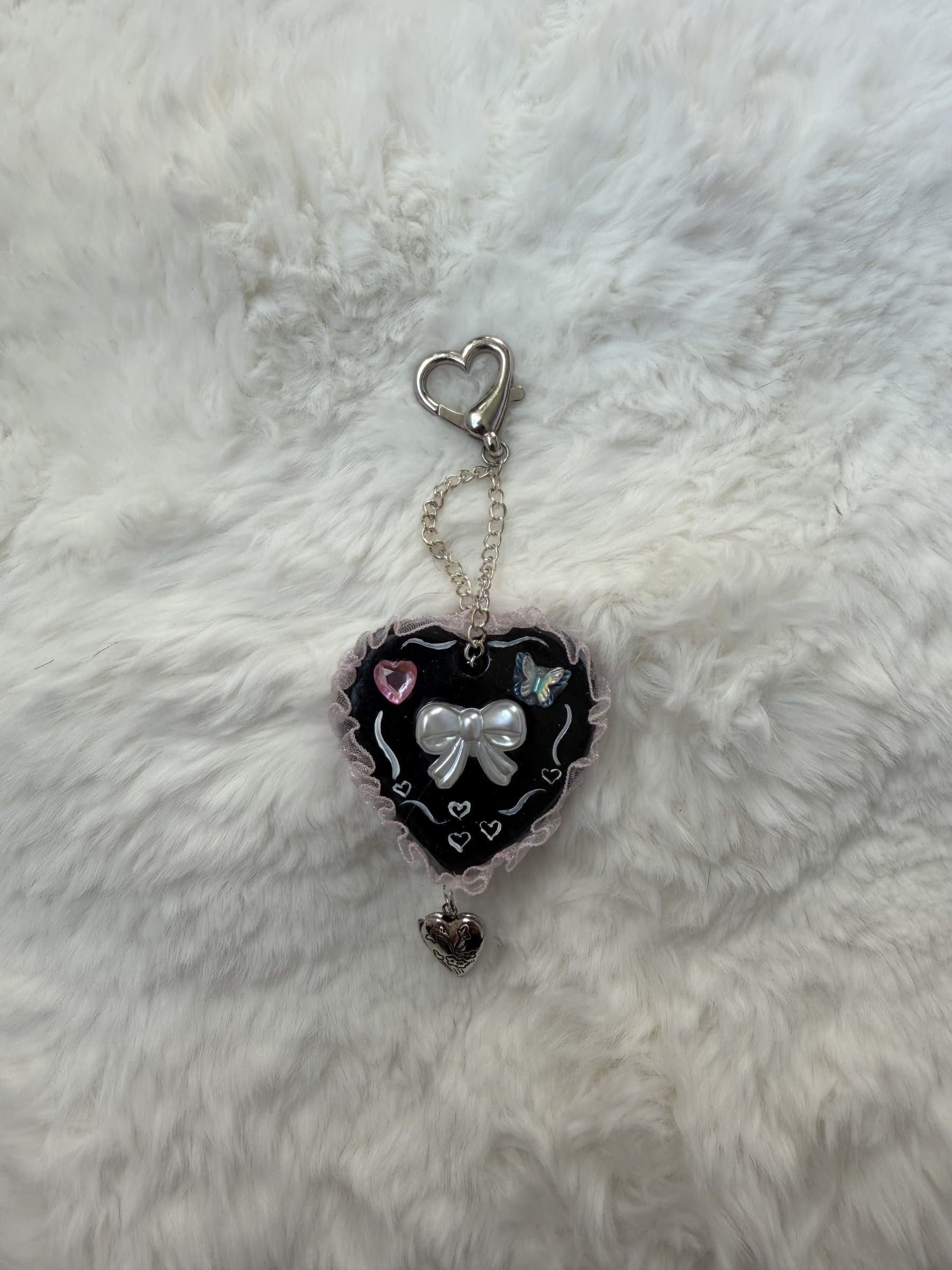 Black clay heart keychain with a pearl bow, pink gem heart, blue butterfly, pink lace trim, and a dangling heart locket