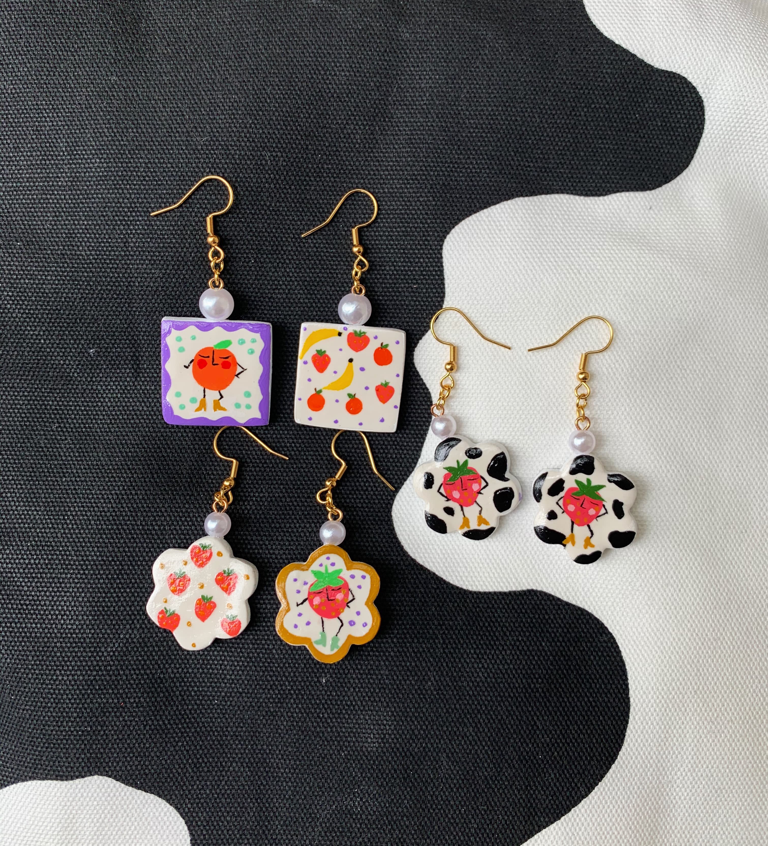 Three pairs of fruit-themed polymer clay earrings on black and white cow-print fabric