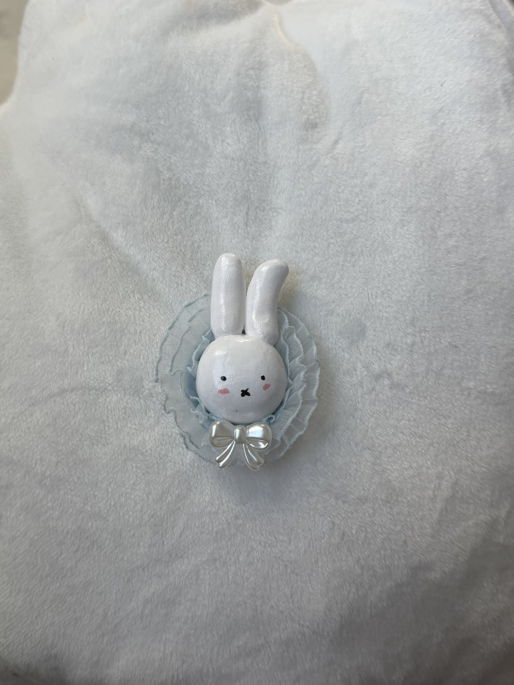 White clay bunny with rosy cheeks in a pale blue lace ruffle with a pearl bow, on a soft blue blanket