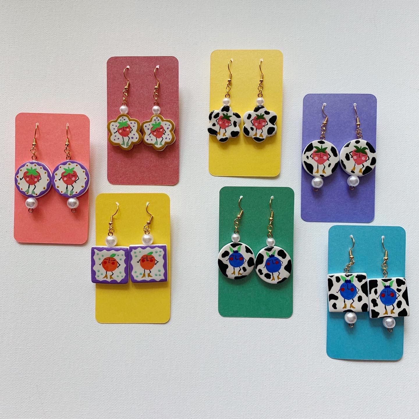 Seven pairs of hand-painted fruit-character polymer clay earrings displayed on colorful cards