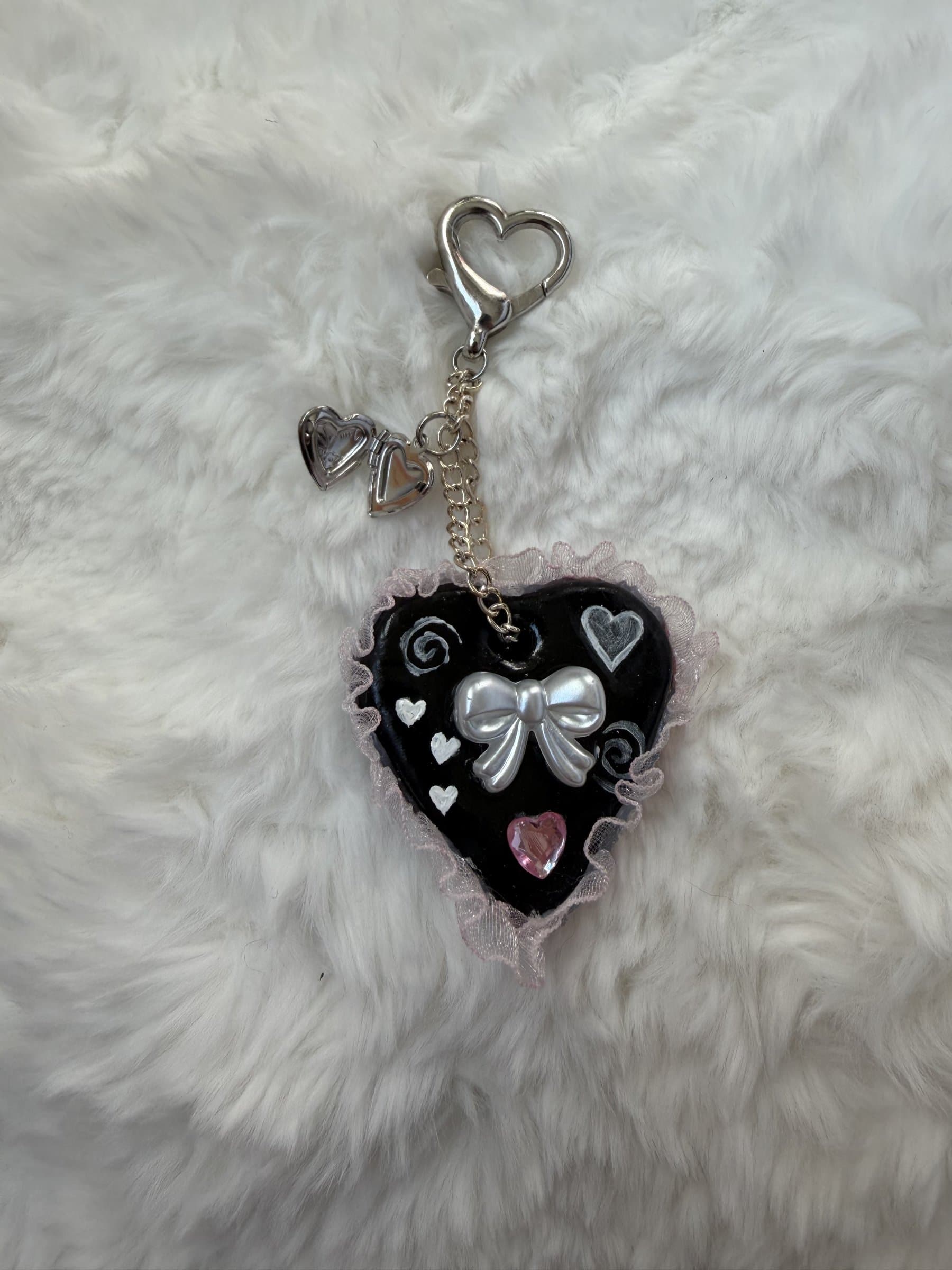 Five black and white clay heart keychains with pearl bows, pink gems, ribbons, and lace ruffles laid out on white faux fur