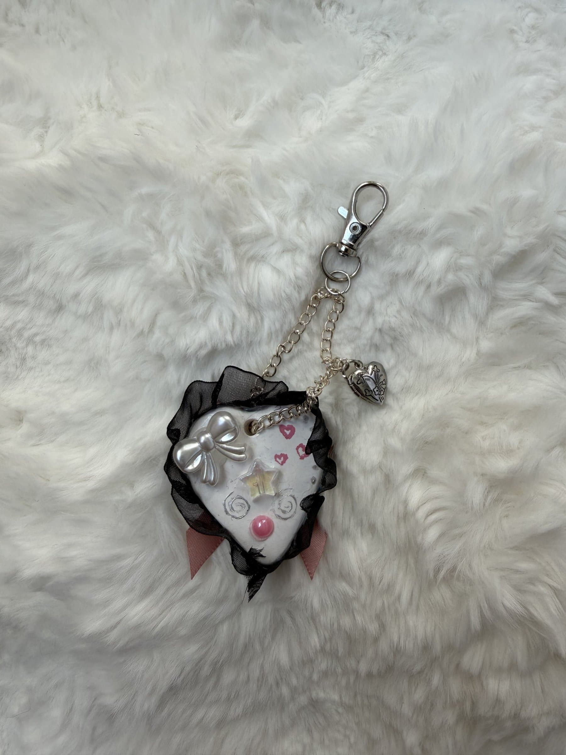 White clay heart keychain trimmed with black ruffle, a dusty pink satin bow, an iridescent star, and a silver chain with an engraved heart locket