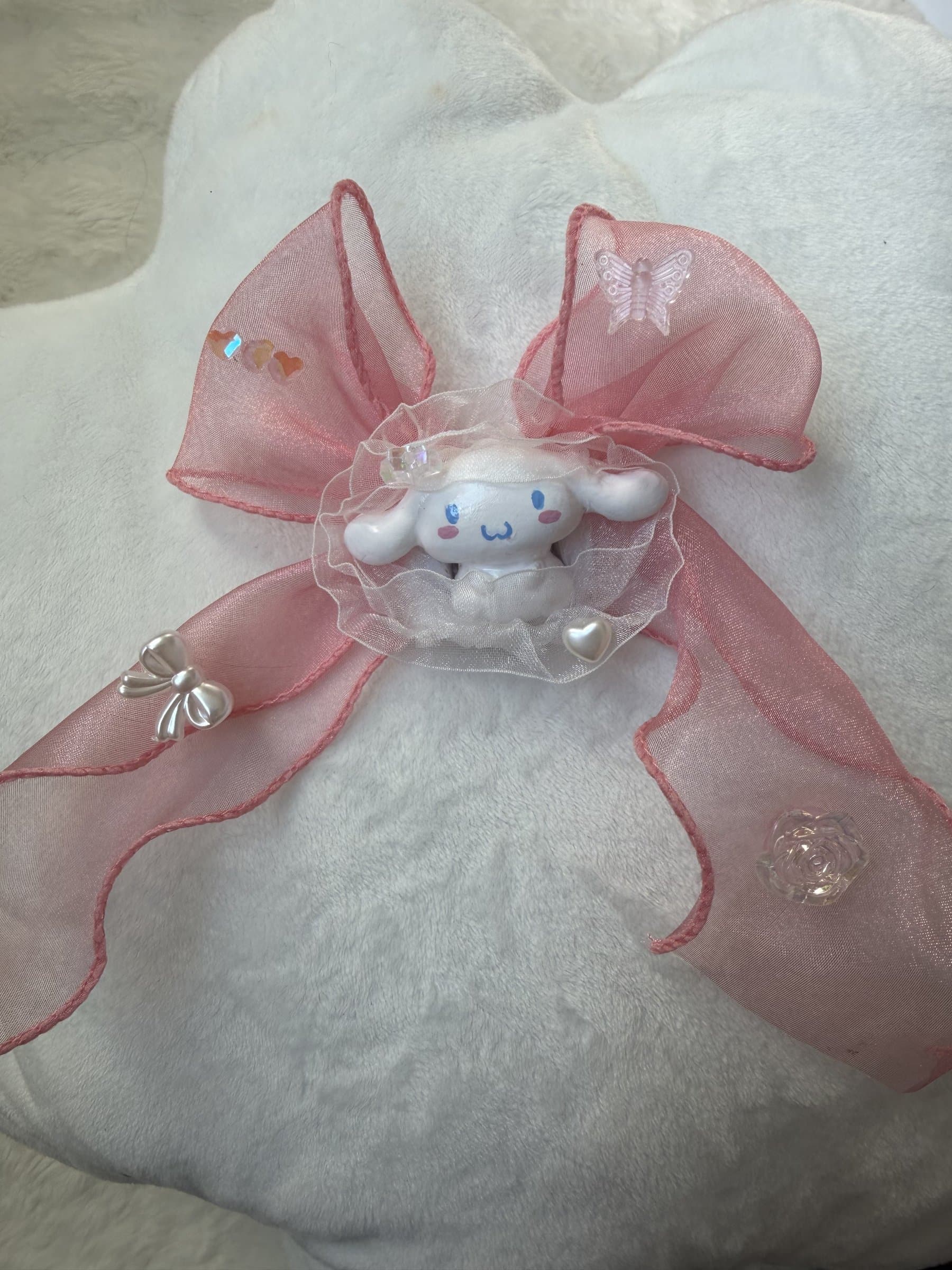 Polymer clay Cinnamoroll puppy resting in a white lace ruffle beneath a large sheer pink bow with rose, butterfly, and heart charms
