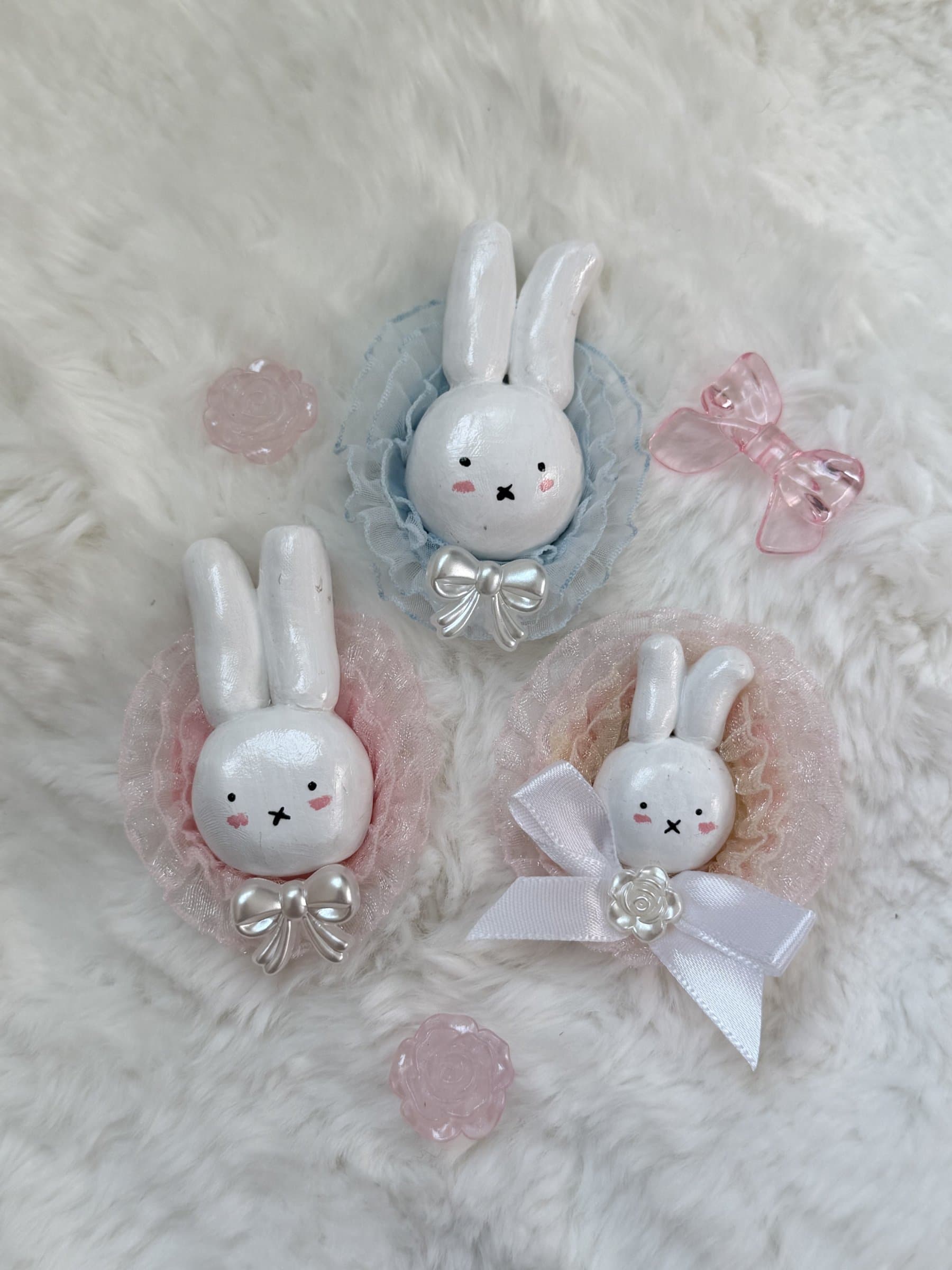 Three clay bunny magnets with ruffled lace collars in blue, pink, and blush, each finished with a pearl bow, beside rose and bow beads